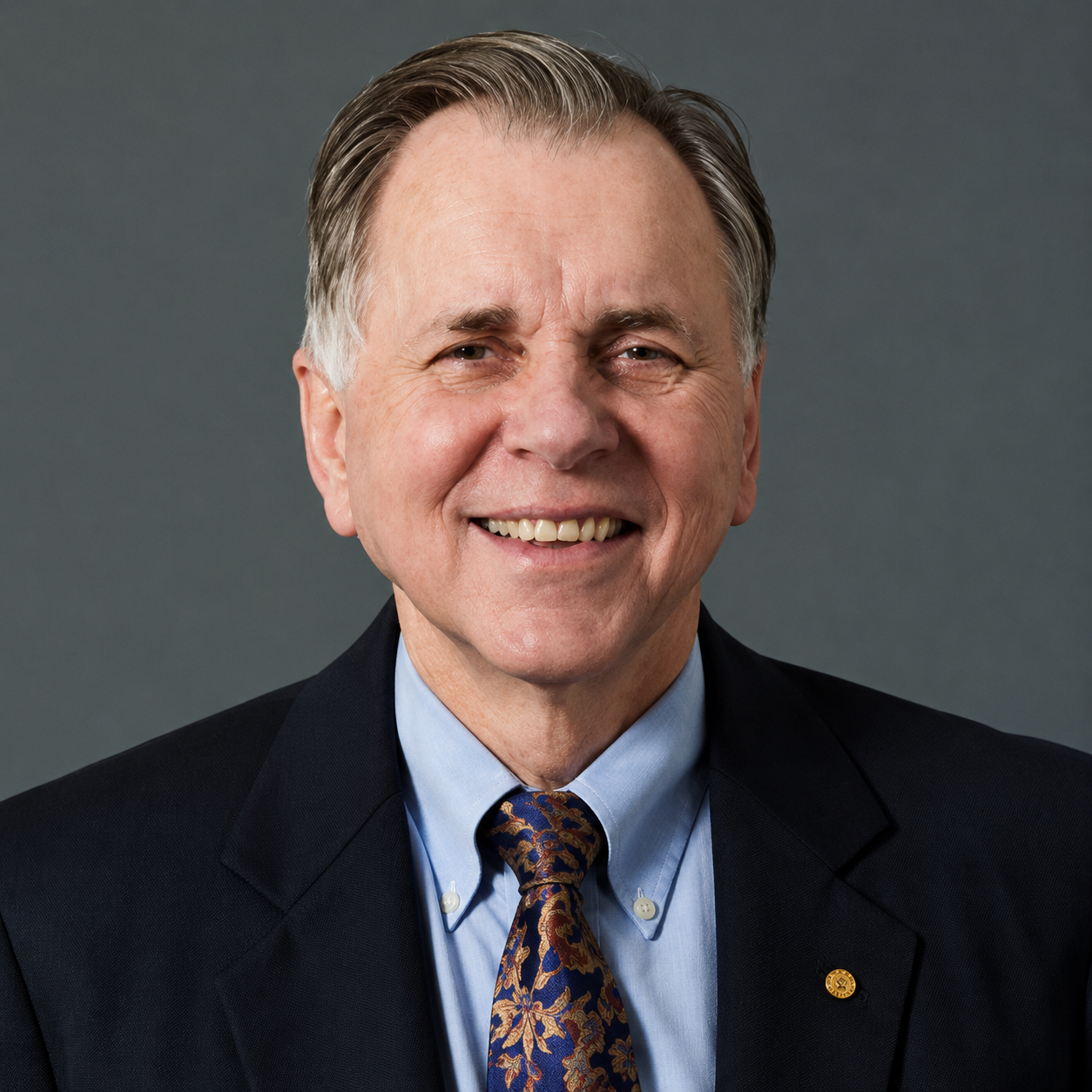 Smiling older man with gray hair wearing a dark suit, blue shirt, and patterned tie against a gray background.